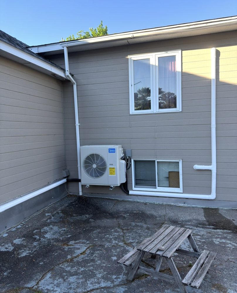 Ductless heat pump outdoor unit mounted on a wall bracket beside a basement window, with white line covers